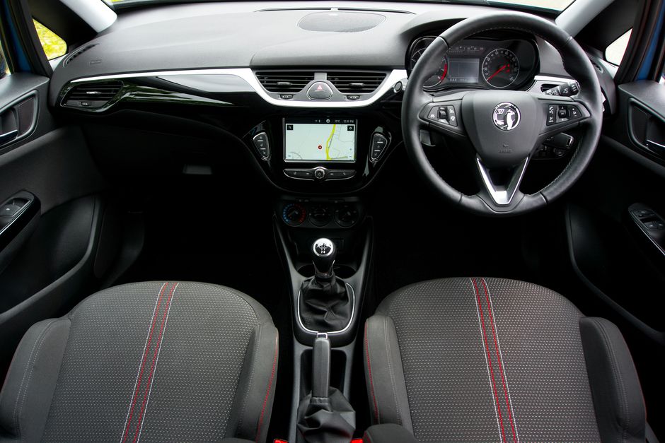 The interior view of a Vauxhall vehicle showing the dashboard, steering wheel, and front seats. The dashboard features a central touchscreen displaying a navigation map, with climate control dials and buttons situated below it. The steering wheel, branded with the Vauxhall logo, has mounted controls on each side. The front seats are upholstered in dark fabric with red stitching and contrasting red and gray patterning running down the center. The gear shift lever is located between the seats, with a boot covering its base, and additional controls and storage compartments are visible around the central console. The vehicle appears to be parked, and the image is captured from the rear-facing perspective inside the car, highlighting the layout and features of the interior, which relates to the context of packing and moving as part of a home relocation process.