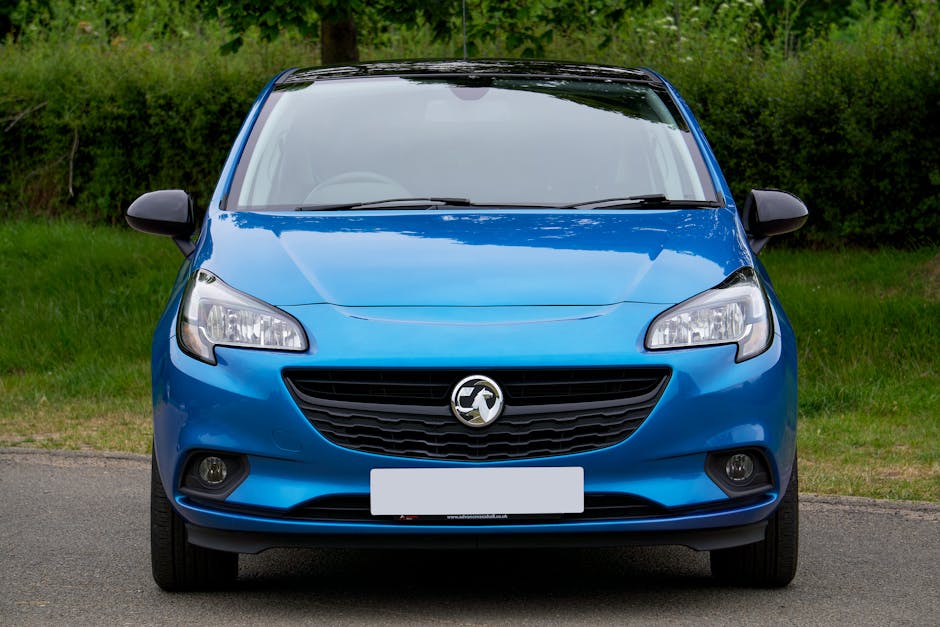 A front view of a blue Vauxhall hatchback parked on a paved driveway with a grassy verge and green trees in the background, featuring clear headlights, a black grille with the Vauxhall emblem, and side mirrors extended. The vehicle is positioned centrally, ready for a home relocation or furniture transport process, with natural daylight illuminating the scene. The image captures the exterior of the car in a neutral setting, associated with moving and delivery services provided by Man With a Van Vauxhall, emphasizing the importance of proper vehicle use during packing and loading operations in the context of delayed lift scenarios on moving day.