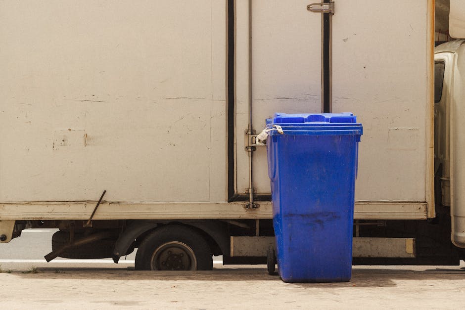 A blue recycling waste bin is positioned outside on a pavement near the rear of a white panel van used for house removals, with the van's sliding door partially visible and slightly open, revealing a glimpse of cardboard boxes and packing materials inside. The bin is standing upright on its wheels, secured by a plastic tie, and is adjacent to the van, which is parked in a residential area. The scene includes a portion of the van's wheel and undercarriage beneath the cargo area, and the environment suggests an exterior loading or unloading process typical of furniture transport and packing during home relocation. The backdrop features a neutral wall and concrete ground, with natural daylight illuminating the scene, emphasizing the loading process related to removal services by Man With a Van Vauxhall.