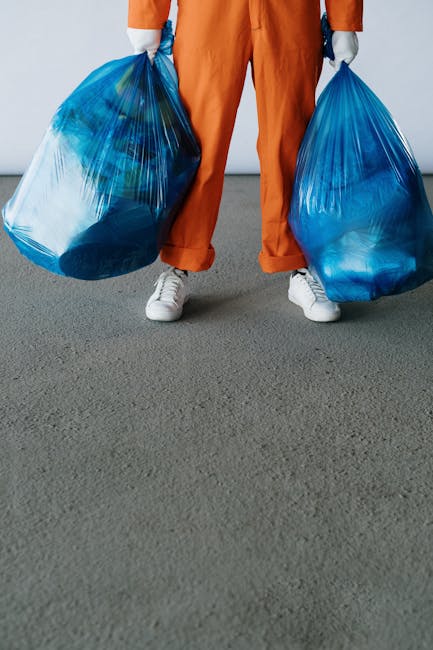 A person wearing orange work trousers, white sneakers, and grey gloves is standing indoors on a grey carpeted floor, holding two large blue plastic bags filled with waste or recyclable materials, one in each hand. Part of their torso and legs are visible, with the bags appearing full and slightly bulging. Behind the individual, a plain white wall and an open doorway are visible, leading to what appears to be a storage or moving area. The scene suggests preparation for waste disposal or packing related to a household move, in line with house removals and packing processes supported by Man With a Van Vauxhall. The environment indicates an interior setting where loading or unloading might be taking place during a furniture transport or home relocation process.