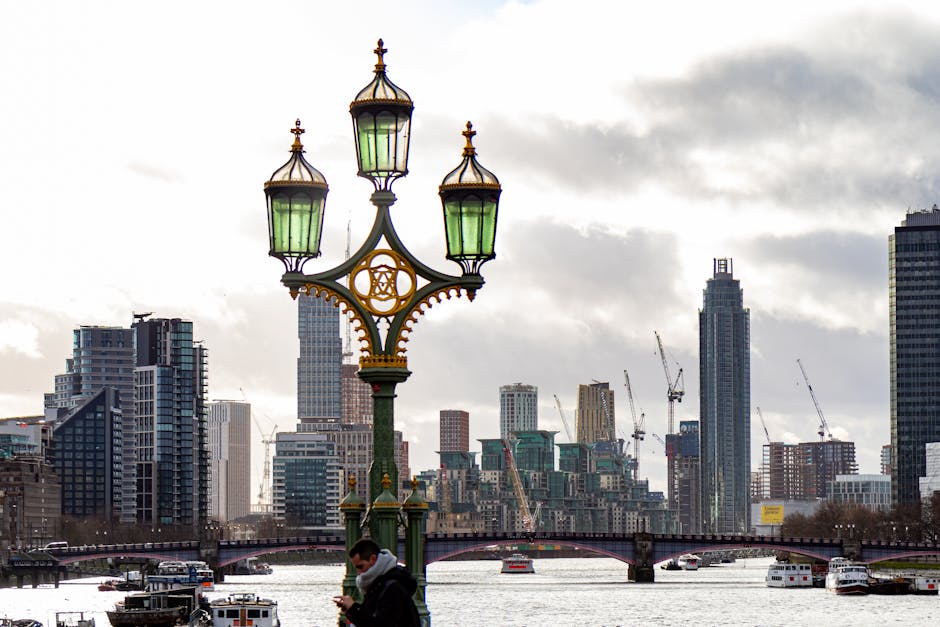 A close-up view of an ornate vintage street lamp with three green-glass lanterns, situated on a paved walkway along the River Thames in Vauxhall, London. In the background, there are modern high-rise buildings under construction, with cranes visible among the skyline, and a few boats on the river. The sky is partly cloudy, casting diffused light over the scene. This setting reflects an urban environment where house removals and furniture transport could take place during the process of relocating homes or offices nearby, with Man With a Van Vauxhall providing comprehensive packing and loading services for property transfers in the SW8 area.