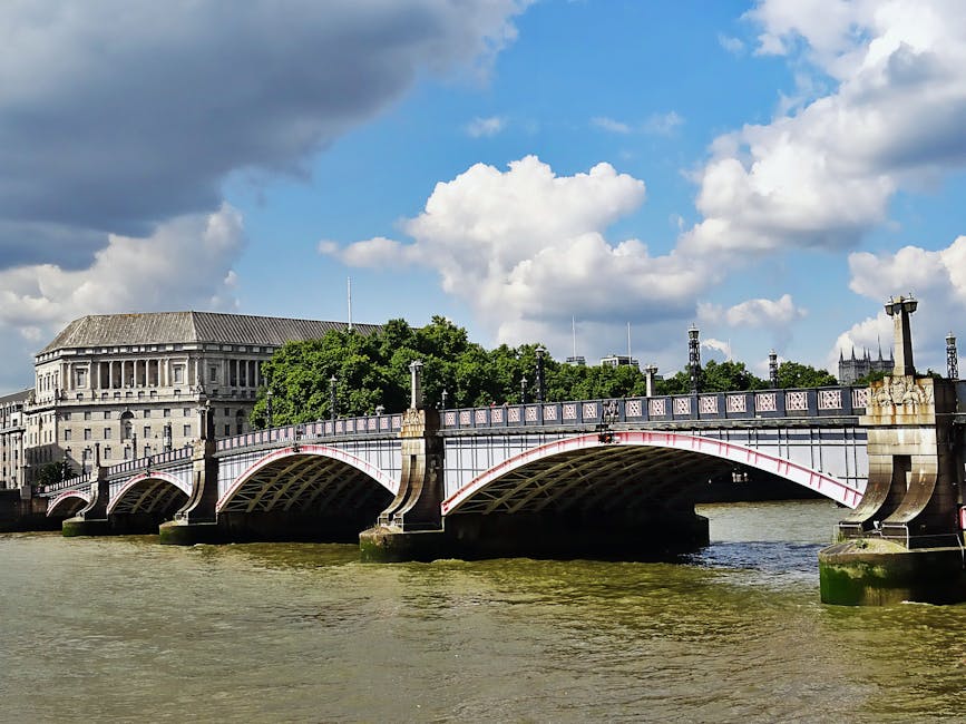 A low-angle view of a historic bridge over the River Thames in London, featuring arched supports and a decorative iron railing on top. Behind the bridge, there is a large, classical-style building with columns and a sloped roof. To the right, a section of a Gothic cathedral with spires is visible in the distance. The sky above is partly cloudy with a mix of dark and white clouds, casting shadows on the water below. The river appears calm with slight ripples. In the foreground, the riverbank is not visible. This scene reflects a typical London landscape, relevant to house removals and relocation logistics in the area, with the bridge and river as part of the urban environment used during furniture transport or home relocation processes managed by companies such as Man With a Van Vauxhall.