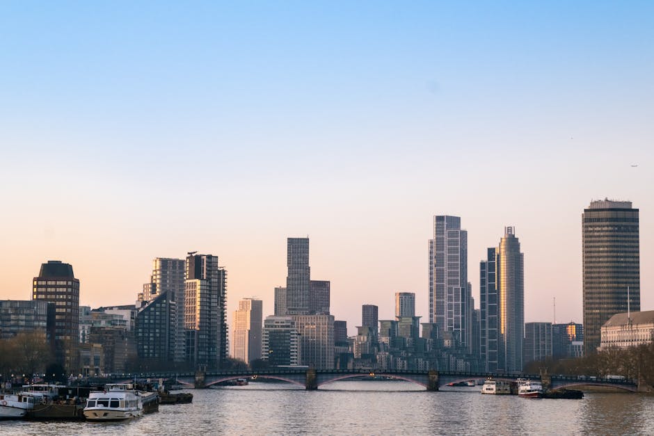 A wide-angle view of the London skyline along the river Thames during the daytime, featuring tall modern skyscrapers and office buildings, with some glass facades reflecting the sky. In the foreground, a calm waterway contains several moored boats and barges, with a small bridge spanning the river. The scene is illuminated by natural light, highlighting the architectural details of the buildings. This urban landscape aligns with house removals and furniture transport, illustrating a typical city environment where home relocation and loading processes may take place, supporting services like those provided by Man With a Van Vauxhall in busy, tight-space moving situations.