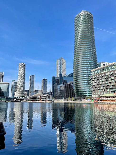 A wide-angle view of the London skyline along the river Thames during the daytime, featuring tall modern skyscrapers and office buildings, with some glass facades reflecting the sky. In the foreground, a calm waterway contains several moored boats and barges, with a small bridge spanning the river. The scene is illuminated by natural light, highlighting the architectural details of the buildings. This urban landscape aligns with house removals and furniture transport, illustrating a typical city environment where home relocation and loading processes may take place, supporting services like those provided by Man With a Van Vauxhall in busy, tight-space moving situations.
