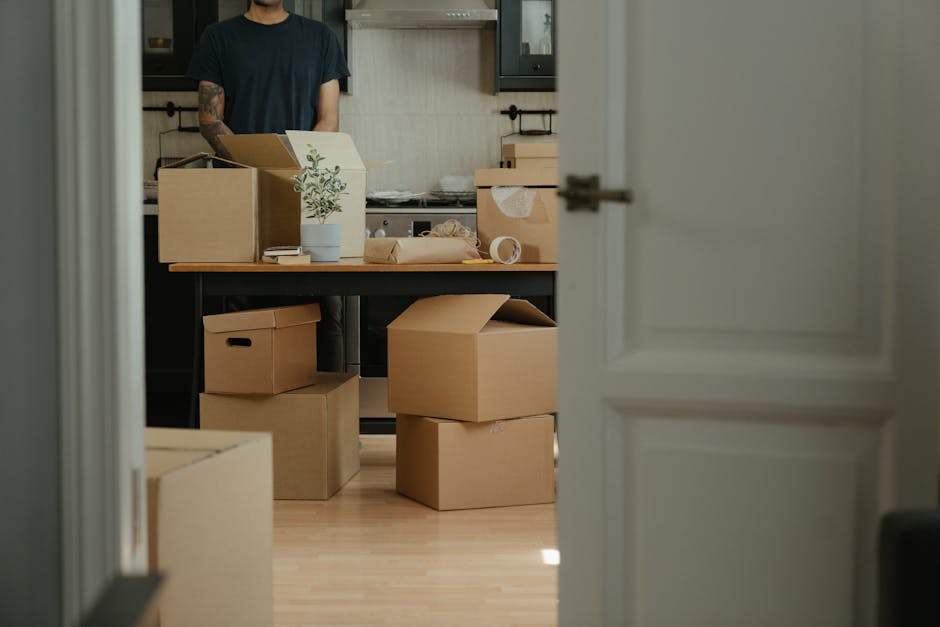 View through an open doorway into a kitchen or living area, where moving boxes made of brown cardboard are stacked on a wooden floor and placed on a black table. Some boxes are closed, while others are partially open or sealed with brown packing tape. On the table, there is a white ceramic pot with a small green plant, and packing materials such as bubble wrap and a roll of packing tape are visible. A person wearing a dark t-shirt, partially visible from the waist up, stands behind the boxes, suggesting the process of packing or preparing for a home relocation. The room features dark kitchen cabinets and a neutral wall finish. This scene depicts the packing and loading process involved in a house removals service by Man With a Van Vauxhall during a move out of a flat on South Lambeth Road, Vauxhall.