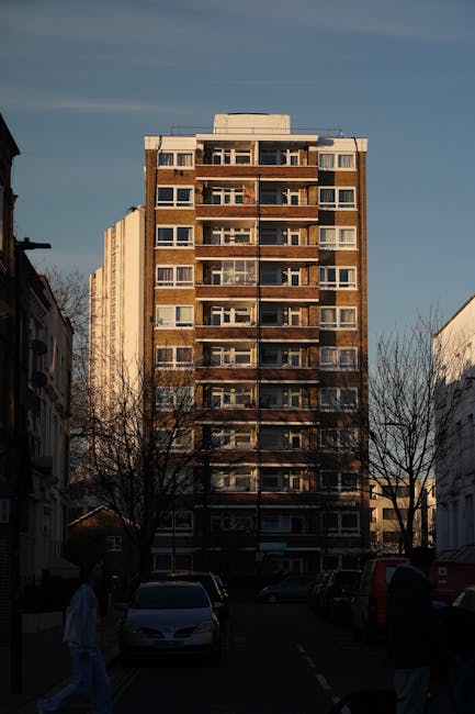 A tall, multi-storey residential building on South Lambeth Road with a rectangular structural design, featuring numerous windows and balconies arranged in vertical columns, under a cloudy sky during daylight. In the foreground, a parking area with several cars is visible, along with two individuals walking on the pavement. The scene is set in an urban environment with leafless trees lining the street, indicating a possible late autumn or winter season. This image relates to the context of house removals and home relocation, emphasizing the building where moving services from Man With a Van Vauxhall might be required for furniture transport, packing, and loading processes during a flat move or residential change.