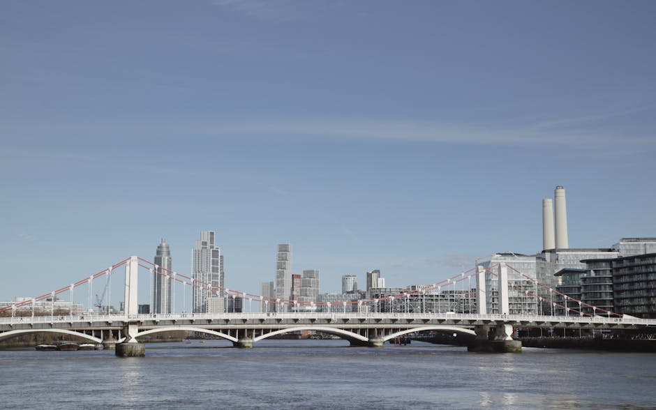 A view of the River Thames with a white suspension bridge in the foreground, supporting traffic and pedestrians. Behind the bridge, the London city skyline features modern high-rise buildings and office towers, some with glass facades reflecting sunlight. To the right, there are two tall smokestacks and a large industrial or commercial building with multiple windows. The scene is captured on a clear day with blue skies and minimal cloud cover, and the river appears calm with gentle ripples. This urban landscape image relates to the area between Nine Elms and Albert Embankment, relevant for home relocation, furniture transport, and moving services offered by Man With a Van Vauxhall.