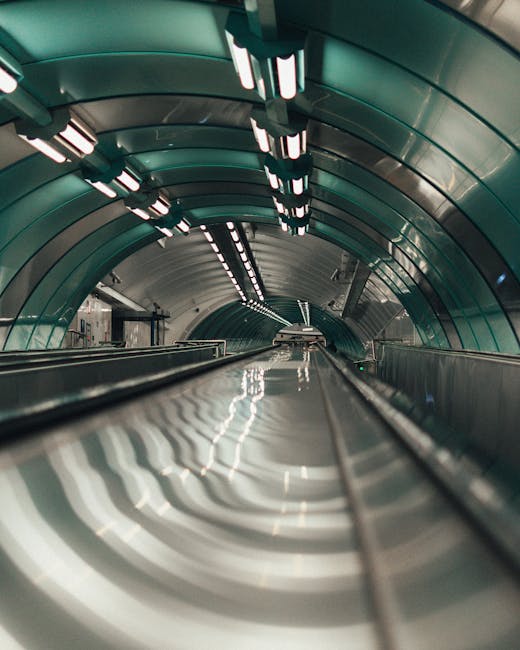 Inside a modern underground tunnel with curved, metallic walls and ceiling panels illuminated by evenly spaced rectangular LED lights. The perspective is from the bottom of an escalator or moving walkway, looking forward along its reflective metal surface towards a vanishing point. The environment appears clean and well-maintained, suggesting a transportation hub or station. This setting illustrates an urban area suitable for home relocation or furniture transport services provided by Man With a Van Vauxhall, linking to efficient moving logistics between Nine Elms and Albert Embankment. The image emphasizes the infrastructure involved in modern city moves, focusing on transportation routes and access points for professional removals and packing and moving operations.