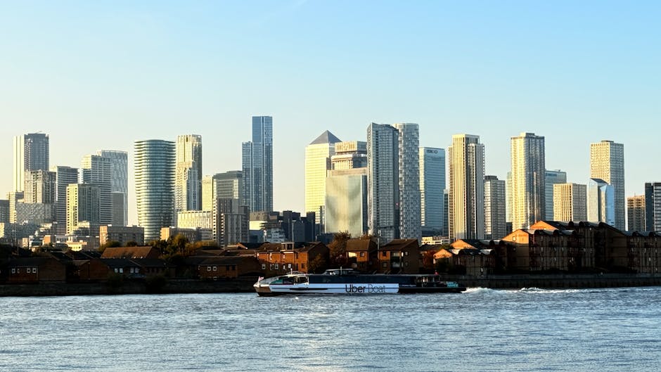 A view of the River Thames with a white suspension bridge in the foreground, supporting traffic and pedestrians. Behind the bridge, the London city skyline features modern high-rise buildings and office towers, some with glass facades reflecting sunlight. To the right, there are two tall smokestacks and a large industrial or commercial building with multiple windows. The scene is captured on a clear day with blue skies and minimal cloud cover, and the river appears calm with gentle ripples. This urban landscape image relates to the area between Nine Elms and Albert Embankment, relevant for home relocation, furniture transport, and moving services offered by Man With a Van Vauxhall.