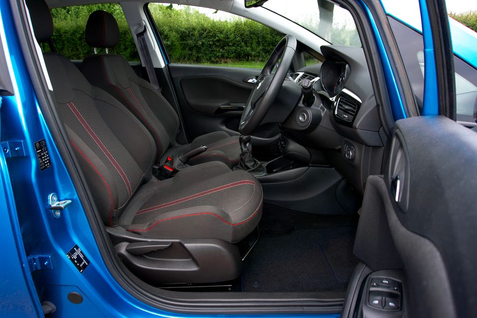 The interior of a small blue van during a home relocation, showing the front passenger and driver seats upholstered in dark fabric with red stitching, positioned in a spacious area inside the vehicle. The dashboard includes a steering wheel, controls, and a digital display, with a gear stick and handbrake visible between the seats. The vehicle is parked outside on a paved surface, with a background of greenery and a hedge, suggesting an outdoor loading or parking area. The interior appears clean and organized, indicating preparation for furniture transport or packing and moving activities, as part of a professional removals process carried out by Man With a Van Vauxhall.
