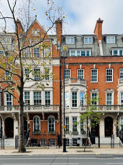 The exterior façade of a Victorian terrace in Vauxhall with red brickwork, white decorative trim, and multiple large windows. The building features ornate stone balconies and a turret-like corner with detailed stonework. In the foreground, a paved sidewalk runs parallel to the street, with a leafless tree and a black lamppost visible. The sky overhead is partly cloudy with patches of blue. This scene represents typical location settings where house removals and furniture transport services by Man With a Van Vauxhall may be performed, illustrating the urban environment involved in relocation logistics, including loading or unloading processes near residential properties.