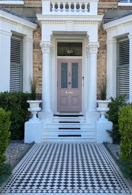 The image shows the entrance to a property located in a Victorian terrace, with a central pastel purple door featuring a glass panel, a brass mail slot, and a door handle. The doorway is framed by two large, white decorative columns with ornate capitals, supporting an entablature with detailed molding. Two symmetrical shrubs in white planters sit on either side of the steps leading up to the door. The steps are white stone with three black strips on each step, and the area in front of the steps is paved with a black and white checkered tile pattern. The entrance is flanked by two windows with white shutters, and above the door is a transom window with the number '50' visible in the background through the glass panel. The façade features a mix of brickwork and white decorative elements, with green bushes lining the sides of the paved walkway, suggesting a neat and well-maintained property. This setting is part of a home relocation or moving process, possibly involving the careful packing, lifting, and transport of household items, as implied by the context of expert removals at Man With a Van Vauxhall.