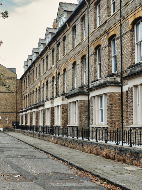 The exterior façade of a Victorian terrace in Vauxhall with red brickwork, white decorative trim, and multiple large windows. The building features ornate stone balconies and a turret-like corner with detailed stonework. In the foreground, a paved sidewalk runs parallel to the street, with a leafless tree and a black lamppost visible. The sky overhead is partly cloudy with patches of blue. This scene represents typical location settings where house removals and furniture transport services by Man With a Van Vauxhall may be performed, illustrating the urban environment involved in relocation logistics, including loading or unloading processes near residential properties.