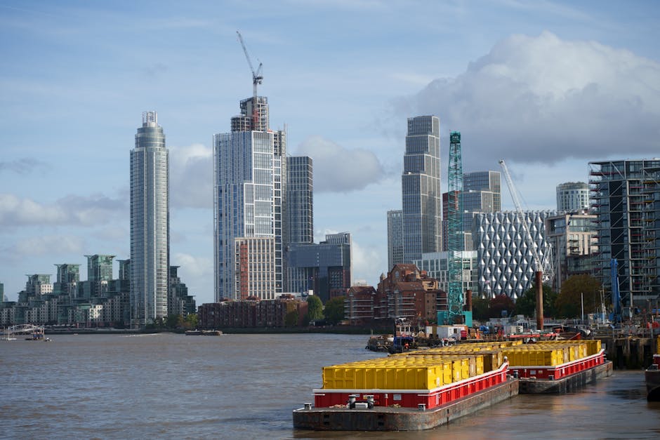 A view of the River Thames in London with modern high-rise buildings and partially constructed skyscrapers in the background. In the foreground, yellow and red floating barriers are positioned along the riverbank, indicating a loading or unloading area. The scene is taken during daylight with partly cloudy skies, and the water reflects the urban skyline. This urban environment highlights the need for efficient home relocation and furniture transport services, like those offered by Man With a Van Vauxhall, which manage packing, loading, and transporting household items during house removals in the city.