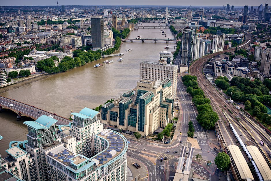 An aerial view of the River Thames flowing through central London, with multiple bridges crossing over it. On the right side, there are high-rise office buildings and modern residential complexes, some with reflective glass facades. The river is dotted with boats and small vessels, and the sky above is partly cloudy. Along the riverbank, a green park area with trees is visible, adjacent to a busy road with cars and buses. To the right, railway tracks run parallel to the road, with a train on the tracks. In the foreground, an area with parking spaces and paved walkways can be seen, with several cars parked. This scene highlights the urban environment involving city infrastructure and development, relevant to relocation and moving logistics as part of house removals or furniture transport projects in London, as documented by Man With a Van Vauxhall.