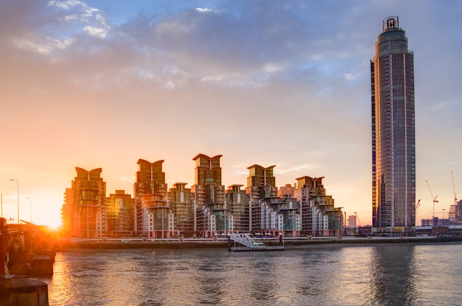 A panoramic view of a city skyline at sunset, featuring a tall, modern skyscraper with a rounded top on the right and a cluster of distinctive, multi-story residential buildings with flat roofs and glass balconies on the left. The buildings are situated along a river, with the water reflecting the warm glow of the setting sun. In the foreground, a few construction cranes are visible, indicating ongoing development, and a small dock with a gangway extends into the water. The sky is partly cloudy with hues of orange, pink, and blue, highlighting the urban landscape, which relates to house removals, furniture transport, and home relocation services provided by companies like Man With a Van Vauxhall.