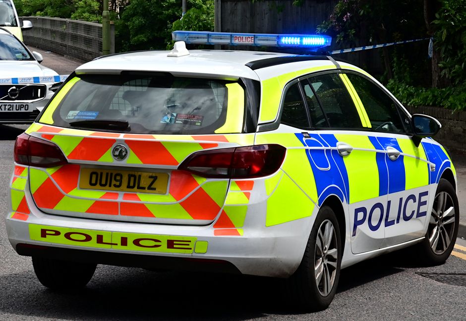 A police car with high-visibility yellow and blue markings and the word 'POLICE' on the side, parked on a street with a police light bar activated on the roof. The vehicle's rear features red and yellow chevron reflective markings, and the registration plate reads 'OU19 DLZ'. In the background, another police vehicle is partially visible, along with greenery and a sidewalk. The scene suggests a police presence during a relocation or movement operation, with the police car positioned on the pavement during daytime, providing a neutral, factual visual context related to traffic or safety enforcement.