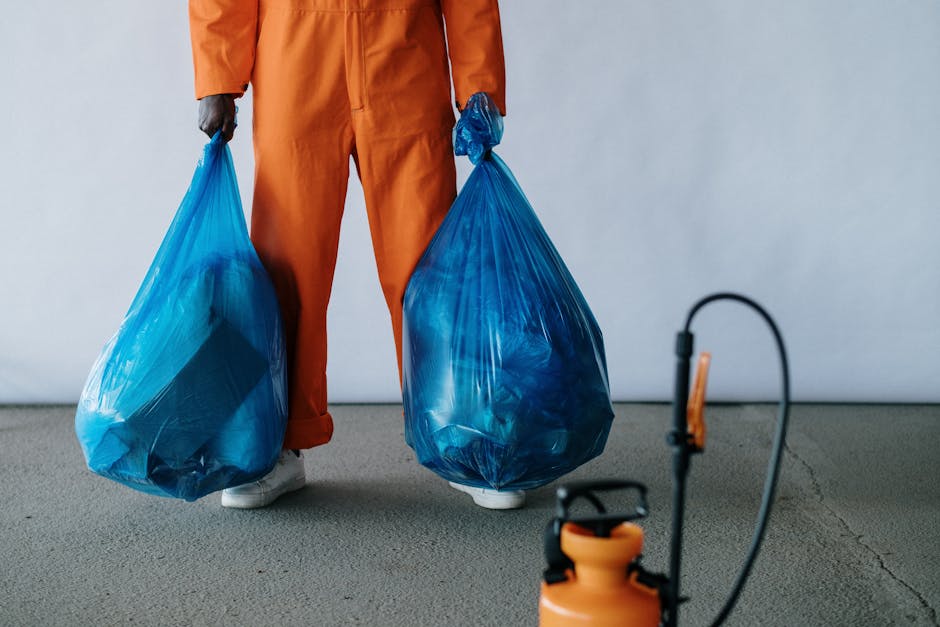 A person dressed in an orange protective suit and white shoes is standing inside an empty room with a plain, light-colored wall and concrete floor. They are holding two large blue plastic garbage bags filled with waste, one in each hand, with the bags partially transparent, showing their contents. The scene appears to capture a waste disposal process as part of a house removal or home relocation, with the individual preparing to load the bags into a van. In the foreground, a small orange portable sprayer or cleaning device is partially visible, suggesting sanitation or cleaning activities associated with moving logistics. The lighting is bright and even, highlighting the materials and action taking place. This image illustrates the process involved in clearing waste during a household move, aligning with the services provided by Man With a Van Vauxhall for efficient removals and responsible waste disposal, particularly under regulations such as those discussed in the 'Waste Disposal Rules in SW8' page.