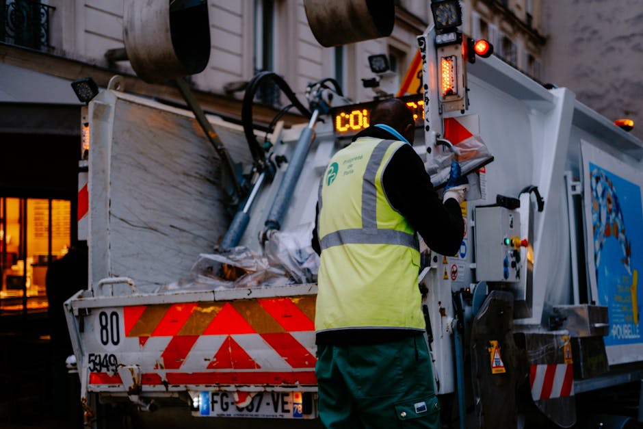 A waste disposal worker wearing a high-visibility vest and dark clothing operates the controls of a large waste collection vehicle with a white and red striped rear section, parked outdoors during evening hours. The worker's back is facing the camera, and they are using gloves to handle the machinery. Inside the vehicle's open container, visible are various discarded packaging materials, including cardboard boxes and plastic wrapping, along with some wooden or metal debris. The vehicle features illuminated warning lights and digital displays on its rear, indicating operational status. The surrounding environment includes a building with warm indoor lighting and large windows in the background, suggesting an urban setting. This image exemplifies the waste collection process typical during home relocations or packing and moving activities, as performed by professional removal services such as Man With a Van Vauxhall.
